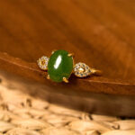 Natural green jade prosperity ring with gold-toned metal band and diamond accents, displayed on wooden tray with rattan mat background