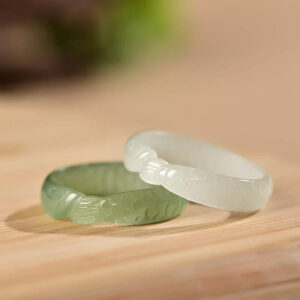 Two handcrafted Hetian jade koi fish rings displayed side by side showing white jade and cyan jade varieties with carved fish details on light wooden surface