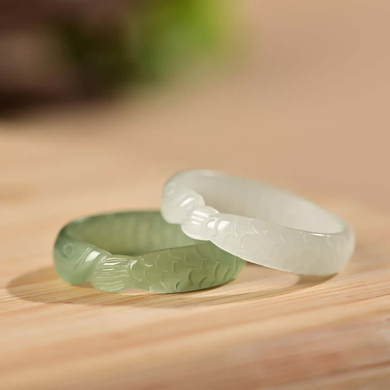 Two handcrafted Hetian jade koi fish rings displayed side by side showing white jade and cyan jade varieties with carved fish details on light wooden surface