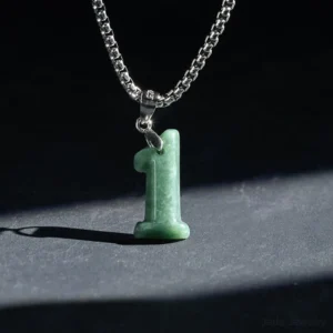 Angel number 1 jade pendant necklace close-up detail, deep green natural jade charm with square link titanium steel chain on dark matte stone background, showing handcrafted pendant attachment