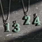 Multiple angel number jade pendant necklaces flat lay display, featuring styles 0-4 with natural green jade charms and premium titanium steel square-link chains on dark stone surface, showing authentic jade stone variations