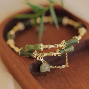 Overhead view of bamboo jade luck bracelet on wooden surface with fresh green bamboo leaves, showcasing jade bell pendant and gold hardware design