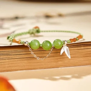 Sterling silver bamboo leaf charm jade beaded bracelet flat lay on vintage book showing green hetian jade beads and braided rope