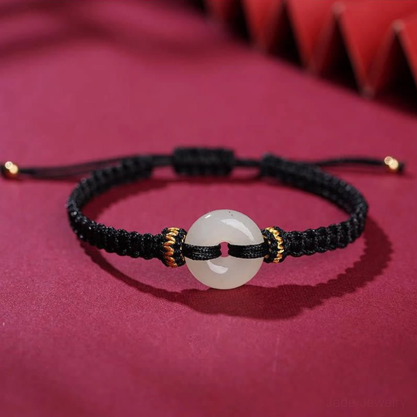 Black braided cord peace buckle jade bracelet with white jade donut pendant and gold accent knots on velvet background