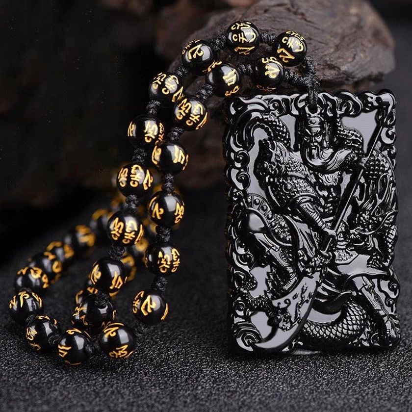 Black obsidian Lord Guan protection pendant with carved Guangong and dragon design, paired with black and gold mantra bead chain on stone display base