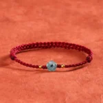 Blue jade flower red string blessing bracelet overhead view with burgundy braided cord and gold spacers on terracotta textile