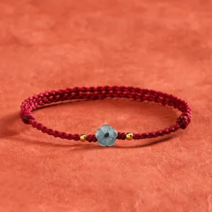 Blue jade flower red string blessing bracelet overhead view with burgundy braided cord and gold spacers on terracotta textile