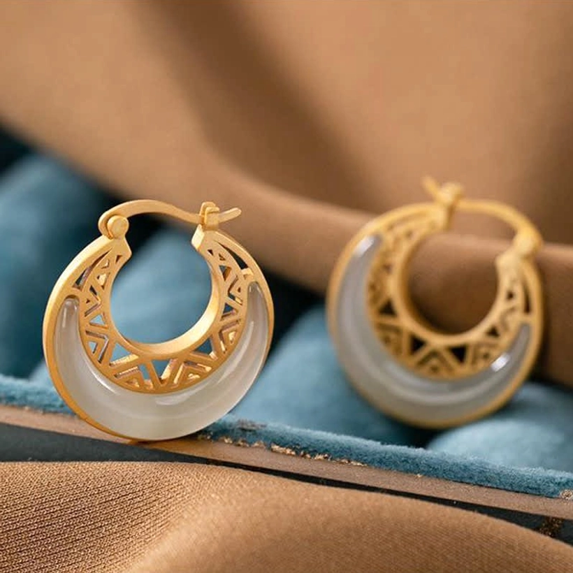 Crescent moon jade drop earrings close-up flat lay on blue velvet tray, milky white jade inlay with matte gold hardware and decorative carved patterns