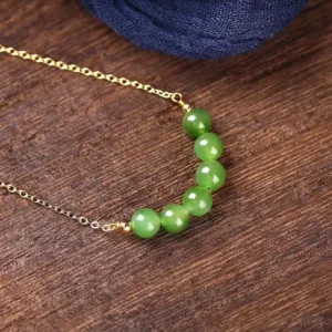 Overhead flat lay of cyan jade beaded necklace showing curved pendant arrangement with seven polished green jade beads on thin 14K gold cable chain against dark wooden background - jade prosperity necklace detail
