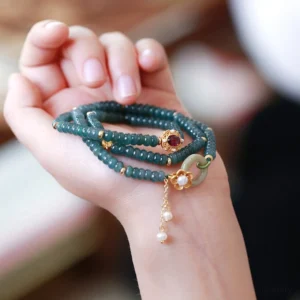 Overhead close-up of stacked cyan jade bracelet with red tourmaline gemstone ornament, pearl pendant, and peace buckle accessory displayed on hand