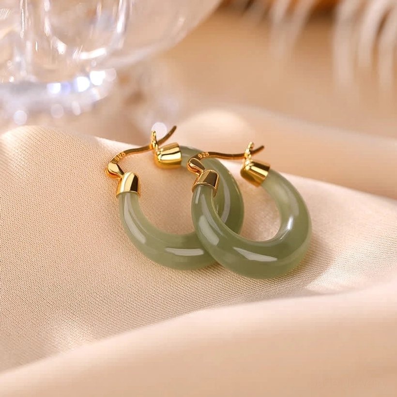 Pair of deep olive green cyan jade drop earrings with gold metal fittings on satin fabric with glassware backdrop, showing rich polished jade finish