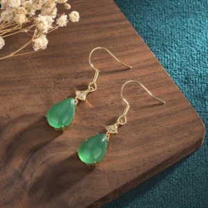 Cyan jade teardrop drop earrings with gold-toned copper hardware on wooden tray with dried flowers and teal fabric, showing overhead oblique view of the elegant green jade stones with zircon accents