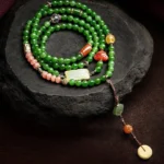 Overhead view of 108 cyan jade mala beads bracelet with laughing Buddha red agate head and hetian jade ruyi charm displayed on dark stone