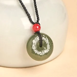 Cyan jade peace buckle necklace featuring dark green jade pendant with carved cloud details, displayed on white ceramic vessel with black braided cord and red accent bead