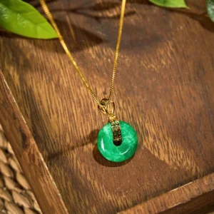 Handcrafted cyan jade peace buckle pendant with delicate gold chain on dark wooden tray, surrounded by fresh green leaves