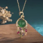 Cyan jade peacock pendant necklace with gold chain and multi-color gemstone inlay, front view on wooden display