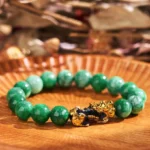 Cyan jade Pi Xiu wealth protection bracelet with gold-plated fortune beast charm, overhead flat lay view showing mixed light and dark green jade beads on wooden display tray