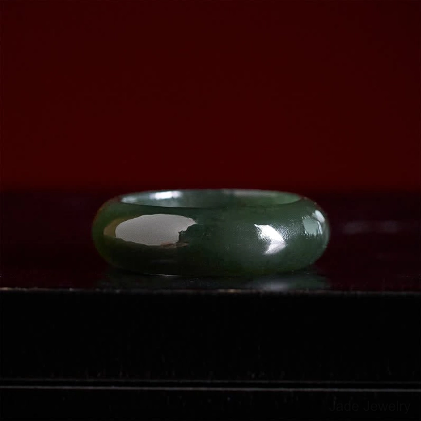Dark cyan jade band ring side profile view on dark maroon surface, displaying ring width and thickness with smooth finish