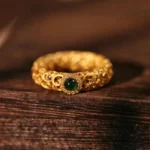 Cyan jade sterling silver filigree ring with gold plating, front top-down view showing intricate hollow carved design with central green jade stone and red accent gems on dark wood surface
