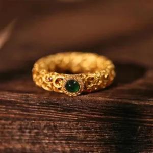Cyan jade sterling silver filigree ring with gold plating, front top-down view showing intricate hollow carved design with central green jade stone and red accent gems on dark wood surface