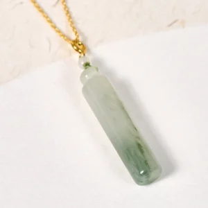 Gold-plated jade cylinder pendant necklace close-up on textured white surface, featuring natural green marbled jade and secure metal clasp