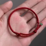 Dark red string jade protection bracelet resting on an open palm, showing the braided cord and light green jade pendant with adjustable knot closure