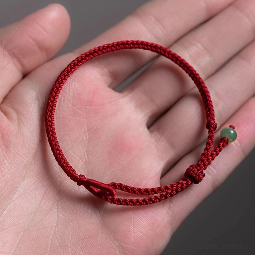 Dark red string jade protection bracelet resting on an open palm, showing the braided cord and light green jade pendant with adjustable knot closure