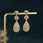 Chinese double happiness jade drop earrings with gold-tone copper studs, enamel cloud motifs, red accent, and pale green teardrop jade pendants displayed on stand