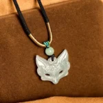 Fox mask jade pendant necklace with black-gold woven rope, light green carved jade fox, overhead front view on brown fabric