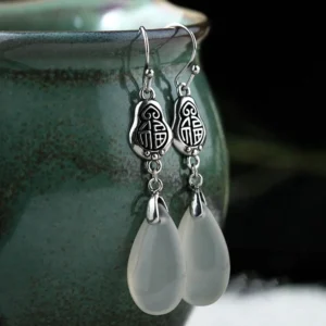 Side profile detail of natural chalcedony teardrop earrings featuring embossed Fu symbol pendant in sterling silver, angled view on ceramic display