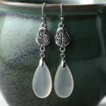 Fu character chalcedony dangle earrings with 925 sterling silver posts displayed on ceramic jar, front close-up view showing embossed Fu symbol and milky white teardrop stone