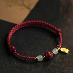 Fu character Hetian jade red string bracelet with carved cinnabar bead and gold Fu charm on dark wooden surface