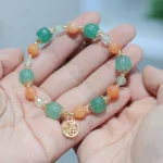 Golden Silk Jade Fu character pumpkin bead bracelet displayed on two palms, showing mixed gemstone beads with gold-toned Fu charm pendant