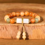 Golden Silk Jade white crystal gourd charm bracelet flat lay on wooden base, warm orange jade beads with white cylindrical charm