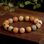 Golden silk jade lotus bracelet overhead view on wooden surface with green foliage, warm tones with black spacer beads and carved lotus charm