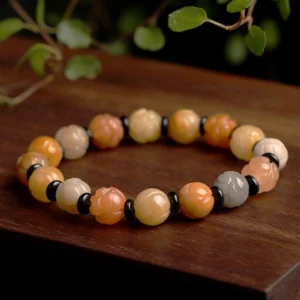 Golden silk jade lotus bracelet overhead view on wooden surface with green foliage, warm tones with black spacer beads and carved lotus charm