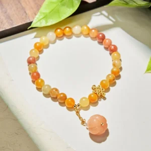 Flat lay top view of Golden Silk Jade pumpkin bracelet showing gradient beads, gold knot charm and pearl pendant on white surface with green leaves