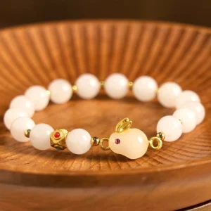 Golden Silk Jade Rabbit Luck Bracelet - Oblique view showing milky white jade beads with gold hardware and red-eyed rabbit charm pendant on wooden tray