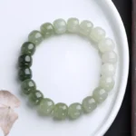 Gradient jade blessing bracelet flat lay showing color transition from dark forest green to milky white barrel beads on white ceramic plate