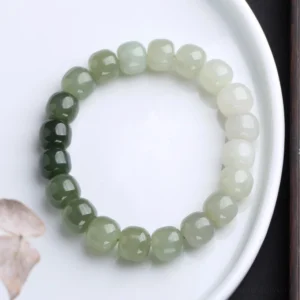 Gradient jade blessing bracelet flat lay showing color transition from dark forest green to milky white barrel beads on white ceramic plate