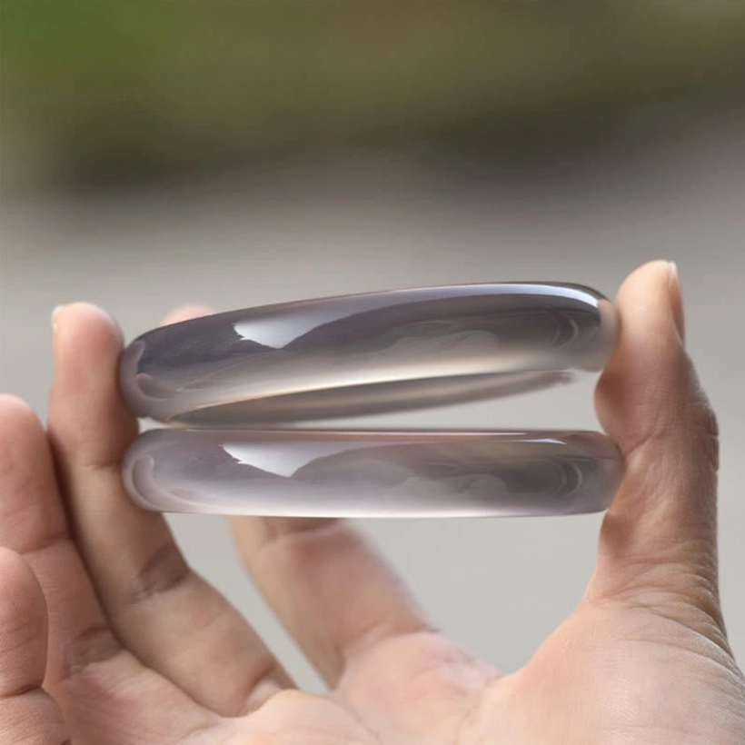 Two gray chalcedony bangles held between thumb and fingers, close-up detail view with soft blurred background