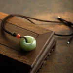 Gray jade peace buckle necklace pendant displayed on wooden tray with braided rope and red accent bead