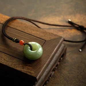 Gray jade peace buckle necklace pendant displayed on wooden tray with braided rope and red accent bead