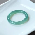 Green chalcedony bangle bracelet displayed on white porcelain plate, showcasing translucent teal green polished stone band