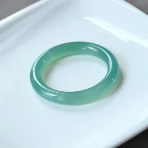 Green chalcedony bangle bracelet displayed on white porcelain plate, showcasing translucent teal green polished stone band