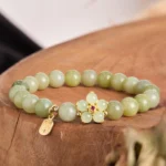 Green jade fu charm bracelet with gold flower pendant displayed on wooden round tray - overhead flat lay view