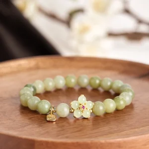 Green jade beaded bracelet with fu character charm and gold flower ornament on wooden tray with floral backdrop