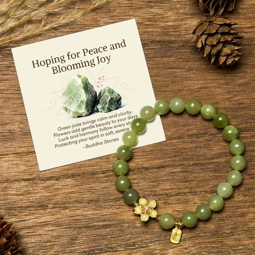 Green jade beaded bracelet with gold flower charm and Fu pendant displayed on wooden table with pinecones and complimentary gift message card