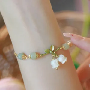 Green jade lily of the valley prosperity bracelet worn on wrist, showing light green jade beads, gold chain, and delicate white lily charms with green leaves