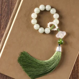 Green jade wrist mala with prosperity tassel displayed on wooden book surface - flat lay view showing pale jade beads and silk tassel